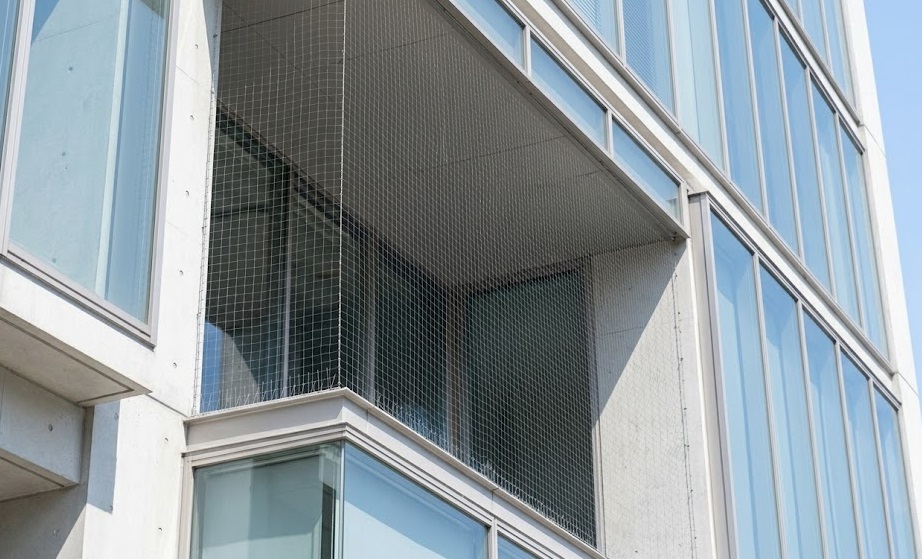福岡市のモダンなガラス張りビルのバルコニーに、極めて細く、下からはほとんど見えない特殊防鳥ネットが施工された後の写真。建物に馴染むデザインで、美観を損なわない solution(Expertise(専門性))の高さと、ハトの侵入を確実に防ぐ高品質な仕上がりを示している。