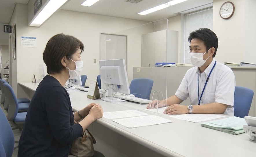 下関市役所の有害鳥獣対策窓口(または一般的な相談カウンター)で、職員(日本人男性)に相談する日本人の女性住民。