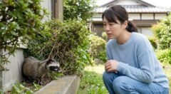 北九州市の住宅庭先で、茂みの陰に隠れるハクビシンを遠くから発見し、驚きながらも安全な距離を保って観察する日本人女性の様子。野生動物との遭遇時の適切な初期対応を描写。