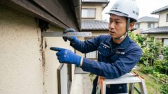 住宅の外壁と軒下のわずかな隙間（1〜2cm）を定規で測定し、コウモリの侵入口であることを示す専門家。