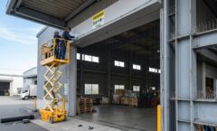 倉庫の大きなシャッター開口部に、プロの業者が高所作業車を使って防鳥ネットを隙間なくピンと張って施工しており、近くのH鋼には防鳥スパイクも設置されている、効果的なハト侵入防止対策の様子。