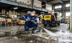 倉庫内で、プロの駆除業者の日本人男性スタッフが、高圧洗浄機を使用して床にこびりついたハトのフンを徹底的に洗浄・掃除している、信頼感のある作業風景。