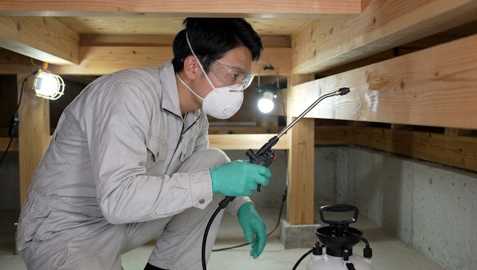 筑紫野市の戸建て住宅の床下で、防護服を着用した誠実な日本人スタッフ（しろあり防除施工士）が、専用の噴霧器を用いて木材と土壌にシロアリ駆除薬剤を散布する「バリア工法」の施工風景。プロフェッショナルな技術と安全管理を示す、E-E-A-T向上のための画像。