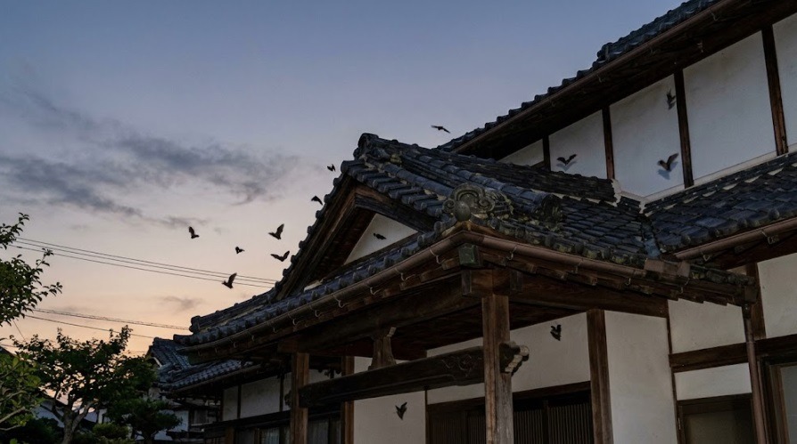 夕方の薄暗い空の下、日本の古い民家の瓦屋根や軒下の隙間から複数の小さなコウモリが飛び回っている様子。