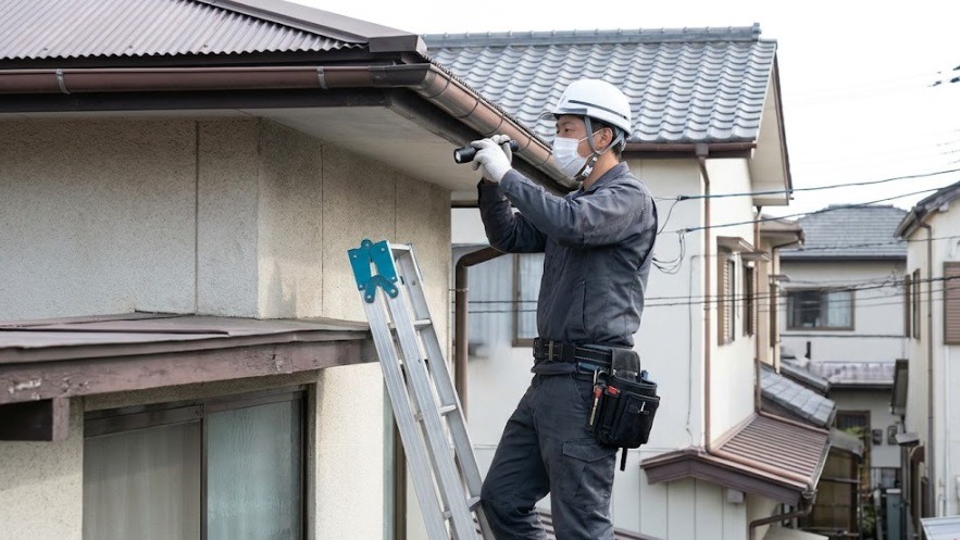 はしご(脚立)に登り、住宅の2階近くの軒下や雨どいの隙間を懐中電灯で細かく調査している、作業服とマスク姿の日本人駆除スタッフ。